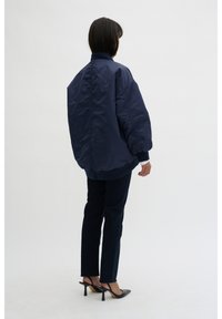 Navy blue bomber jacket with a high collar, oversized silhouette, and elastic cuffs, paired with dark denim pants and black high-heeled shoes.