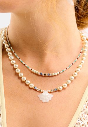 Deux colliers superposés avec de grandes perles rondes, des perles multicolores et un pendentif en forme de coquillage au centre. Accents dorés visibles.