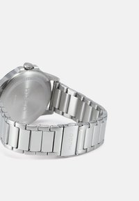 Montre en métal argenté avec un bracelet à maillons, un boîtier rond et une couronne. Dos gravé avec "HUGO." Texture lisse, design moderne.