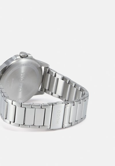 Montre en métal argenté avec un bracelet à maillons, un boîtier rond et une couronne. Dos gravé avec "HUGO." Texture lisse, design moderne.