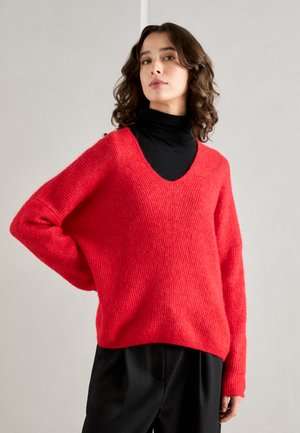 Red, oversized sweater with a deep V-neck, textured knit fabric, and dropped shoulders, paired with a black turtleneck and black trousers.