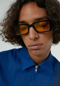 Lunettes de soleil rectangulaires noires avec des verres teintés orange, assorties à une chemise zippée bleue. Textures notables dans les deux matériaux.
