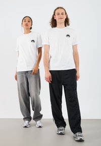 Hvite grafiske t-skjorter med et svart sirkulært design på brystet, kombinert med avslappede grå denim og svarte joggebukser.