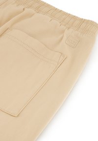 I jogger beige presentano una vita elastica, tasche laterali e una tasca posteriore con un'etichetta logo. Il tessuto ha una texture liscia e morbida.