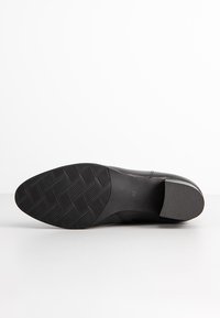 Suola di scarpa in pelle nera con superficie in gomma testurizzata. Presenta un motivo ondulato, bordi lisci e un leggero tacco per stabilità.