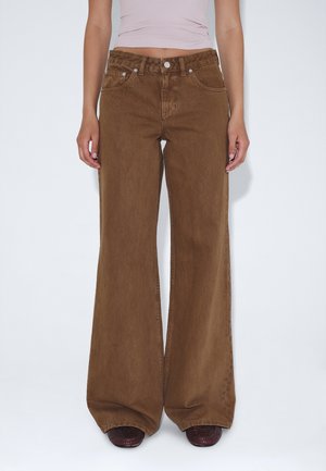 Person iført højtaljede, viddebrune jeans kombineret med en lyserød crop top og mørkebrune sko.