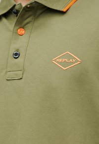 Κοντινή λήψη πράσινου ελιάς πόλο πουκάμισου με πορτοκαλί και μαύρα κουμπιά και πορτοκαλί λογότυπο Replay σε σχήμα ρόμβου στο στήθος.