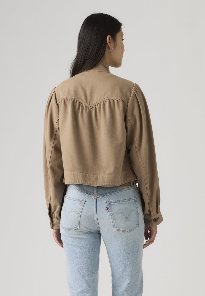 Mujer con cabello largo y oscuro, vestida con una chaqueta marrón con botones y vaqueros azul claro, de pie frente a un fondo liso de color gris claro.