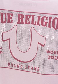 Tissu rose avec un grand texte audacieux "True Religion", des ornements en sequins et un logo fer à cheval stylisé au-dessus de l'inscription "Brand Jeans".
