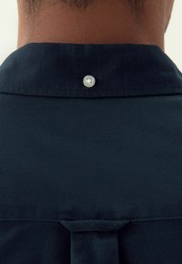 Közeli kép egy személy nyakáról és felső hátáról, aki sötét tengerészkék inget visel, fehér gombbal, ami a gallér alatt középen található.