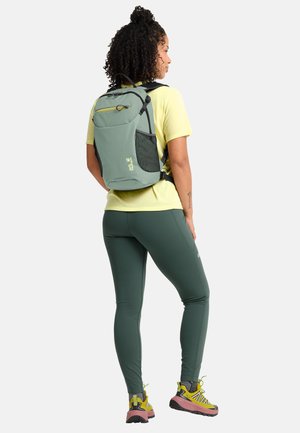 Donna con capelli ricci che indossa uno zaino verde, maglietta gialla, leggings verdi e scarpe da trekking gialle, in piedi davanti a uno sfondo semplice.