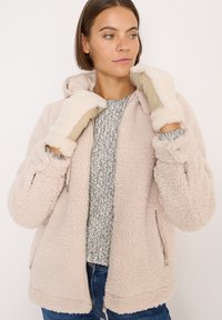 Rosa, fluffy fleecejakke med hætte, udstyret med lynlåslommer og bløde beige handsker. Strikket sweater med tekstur under.