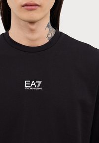 Svart bomull T-skjorte med rund hals og hvit EA7 Emporio Armani-logo i midten. Sleten tekstur, avslappet design.