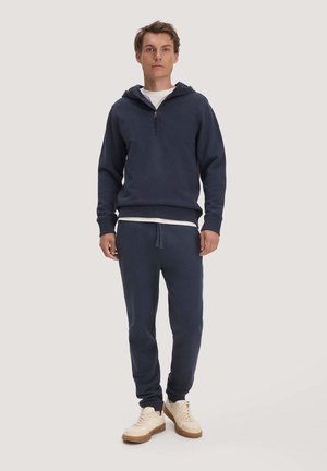 Man staat, draagt een donkerblauwe hoodie met een rits aan de voorkant, bijpassende joggingbroek, wit T-shirt en beige sneakers met rubberen zolen.