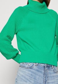 Pull vert en tricot avec un col haut, des manches texturées et un ourlet côtelé, associé à un jean bleu clair. Texture douce et coupe ajustée.