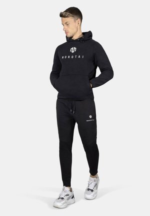 Sort hættetrøje med "Morotai" logo på brystet, parret med matchende sorte joggers med logo detalje. Båret med hvide og grå sneakers.