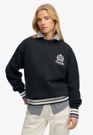 Blondynka w czarnej bluzie Superdry na paskowanej koszuli i kremowych spodniach, stoi z jedną ręką na biodrze.