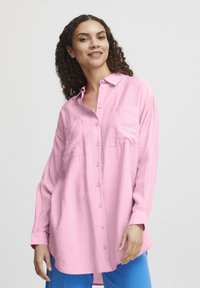 Helles rosa Langarmhemd mit Knopfleiste, zwei Brusttaschen, Kragen und lässiger Passform. Über leuchtend blauen Hosen getragen.