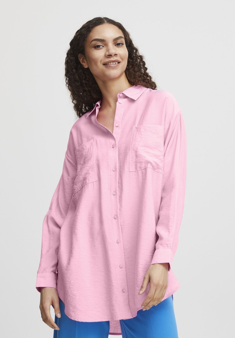 Helles rosa Langarmhemd mit Knopfleiste, zwei Brusttaschen, Kragen und lässiger Passform. Über leuchtend blauen Hosen getragen.