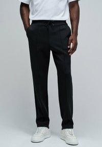 Pantalones deportivos negros con cintura ajustable, corte recto y un sutil detalle en las costuras laterales, combinados con zapatillas blancas.