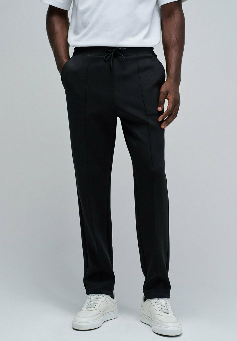 Pantalones deportivos negros con cintura ajustable, corte recto y un sutil detalle en las costuras laterales, combinados con zapatillas blancas.