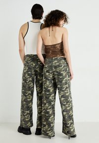 Τα καμουφλάζ cargo παντελόνια διαθέτουν άνετη γραμμή με πολυχρωμο πράσινο σχέδιο. Το μοντέλο στα αριστερά φοράει ένα λευκό tank top, ενώ το μοντέλο στα δεξιά φοράει μια καφέ πλεχτή μπλούζα.