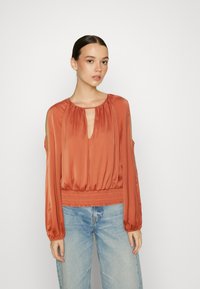 Blusa de seda cor de ferrugem com mangas longas, decote em forma de chave e cintura franzida. Apresenta ombros à mostra e uma textura suave. Combinada com jeans claros.