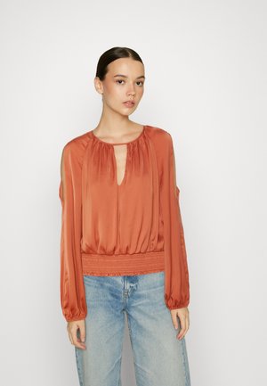 Blusa de seda cor de ferrugem com mangas longas, decote em forma de chave e cintura franzida. Apresenta ombros à mostra e uma textura suave. Combinada com jeans claros.