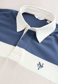 Polo de rugby col avec des rayures horizontales bleu marine, blanc et beige et des initiales brodées "NT" sur la poitrine.