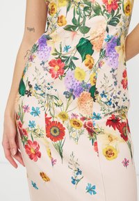 Φλοράλ φόρεμα με κόκκινα, κίτρινα, μωβ και μπλε λουλούδια, φορεμένο από ένα άτομο με τατουάζ στα χέρια που φαίνονται πάνω σε λευκό φόντο.