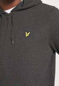 Felpa con zip di colore grigio scuro, realizzata in un tessuto misto cotone. Presenta un logo ricamato di un'aquila gialla sul petto e un cappuccio regolabile con coulisse.