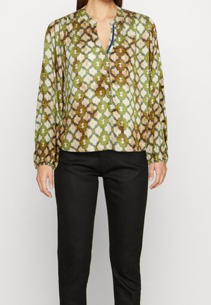 Femme portant un chemisier à manches longues avec des motifs géométriques verts et bruns et un pantalon noir ajusté, debout devant un fond uni.