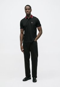 Melnas polo krekls ar sarkaniem akcentiem, īsām piedurknēm un izšūtu logotipu. Kombinēts ar melnām biksēm un melniem apaviem, stāvot pret vienkāršu fona sienu.