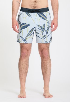 Homme portant un short de bain bleu clair avec un motif de feuilles bleu marine et jaune et un cordon noir, pieds nus sur fond blanc.