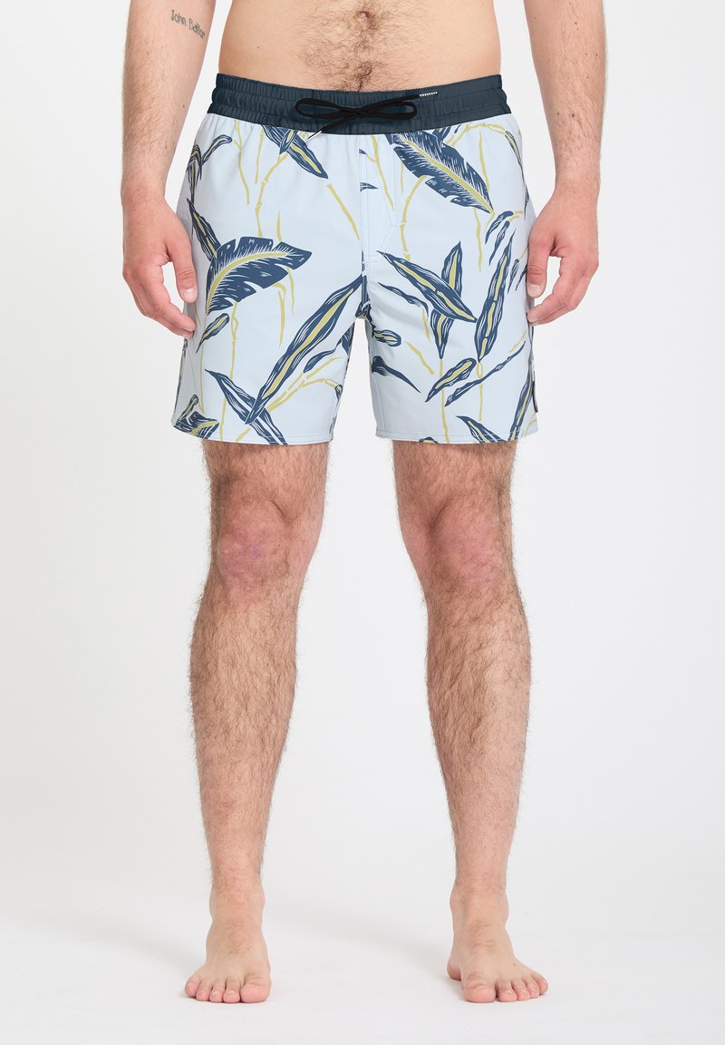Mann trägt hellblaue Badeshorts mit marineblauem und gelbem Blattmuster und schwarzem Kordelzug, barfuß auf weißem Hintergrund.