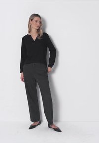 Donna con lunghi capelli biondi che indossa un maglione nero a V, pantaloni grigio scuro e ballerine nere a punta, in piedi contro uno sfondo bianco.