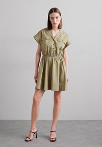 Beige Kleid mit kurzen Ärmeln, Knopfleiste vorne, zwei Brusttaschen, elastischem Bund und ausgestellt geschnittenem Rock. Der Stoff wirkt glatt mit matter Oberfläche.