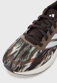 Αθλητικό παπούτσι με υφασμάτινη επιφάνεια με καφέ, πράσινο και μαύρο αφηρημένο καμουφλάζ μοτίβο· διαθέτει καφέ κορδόνια και λευκές λεπτομέρειες.