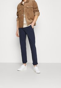 Brun kordjacka med två bröstfickor, randig skjorta under, marinblå chinos och vita sneakers. Uppvikta ärmar på byxorna.