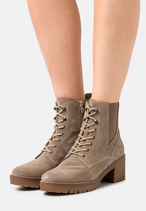 Bottines en suède beige avec un laçage à l'avant, des fermetures éclair sur le côté, des panneaux élastiques et des talons épais, portées sur des jambes nues.