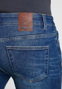 Granatowe dżinsy z denimu z skórzaną łatką na pasie z napisem "TOMMY & SONS". Posiadają przeszycia i tylne kieszenie z lekkim wyblaknięciem.