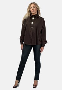 Brązowa, oversize koszula z dużymi białymi guzikami, zestawiona z czarnymi dopasowanymi spodniami i sandałami na obcasie. Gładki materiał, luźny krój, długie rękawy.