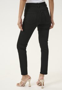 Schwarze High-Waist-Jeans mit slim Fit, die zwei Gesäßtaschen und eine glatte Stofftextur aufweisen. Beigefarbene Absatzsandalen ergänzen den Look.