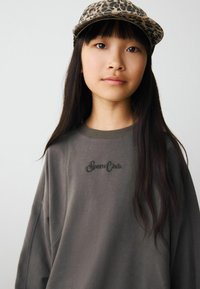 Sweatshirt gris foncé avec un col rond, arborant "Sports Club" brodé en noir. Porté avec une casquette de baseball à imprimé léopard beige.