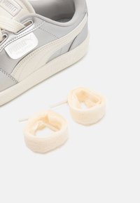 Grå lærersneaker med hvite detaljer og teksturert såle, kombinert med to kremfargede snørebånd med blomsterdesign.