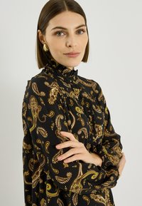 Blusa negra con motivo dorado de paisley, cuello alto con volantes, mangas largas y detalles fruncidos. Tela suave con una silueta fluida.
