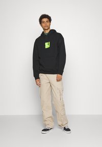 Felpa nera con logo centrale verde, abbinata a pantaloni cargo beige con molte tasche, indossata con sneakers nere e bianche.