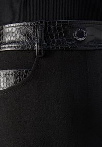 Primo piano di pantaloni neri testurizzati con cintura in pelle di coccodrillo lucida e bordi delle tasche, presenta un bottone metallico rotondo con logo.
