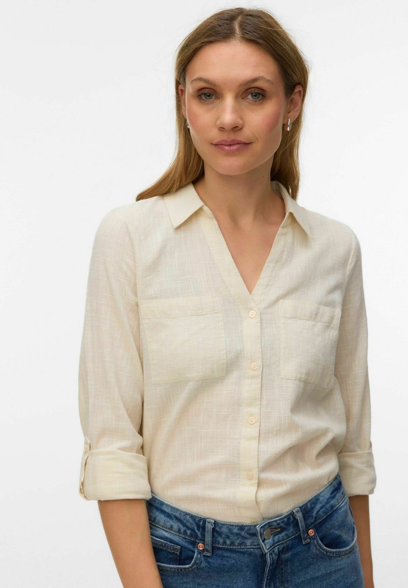 Femme aux cheveux châtain clair portant une chemise crème à boutons avec les manches retroussées et un jean bleu, debout devant un fond blanc uni.