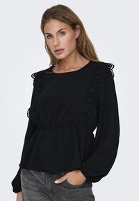 Svart bluse med lange ermer, rund halsutringning og blondeaksenter på skuldrene. Laget av myk, teksturert stoff med peplumstil.